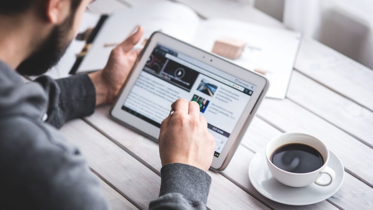 Persoană care citește știri financiare pe o tabletă, la o masă cu o cafea, dimineața
